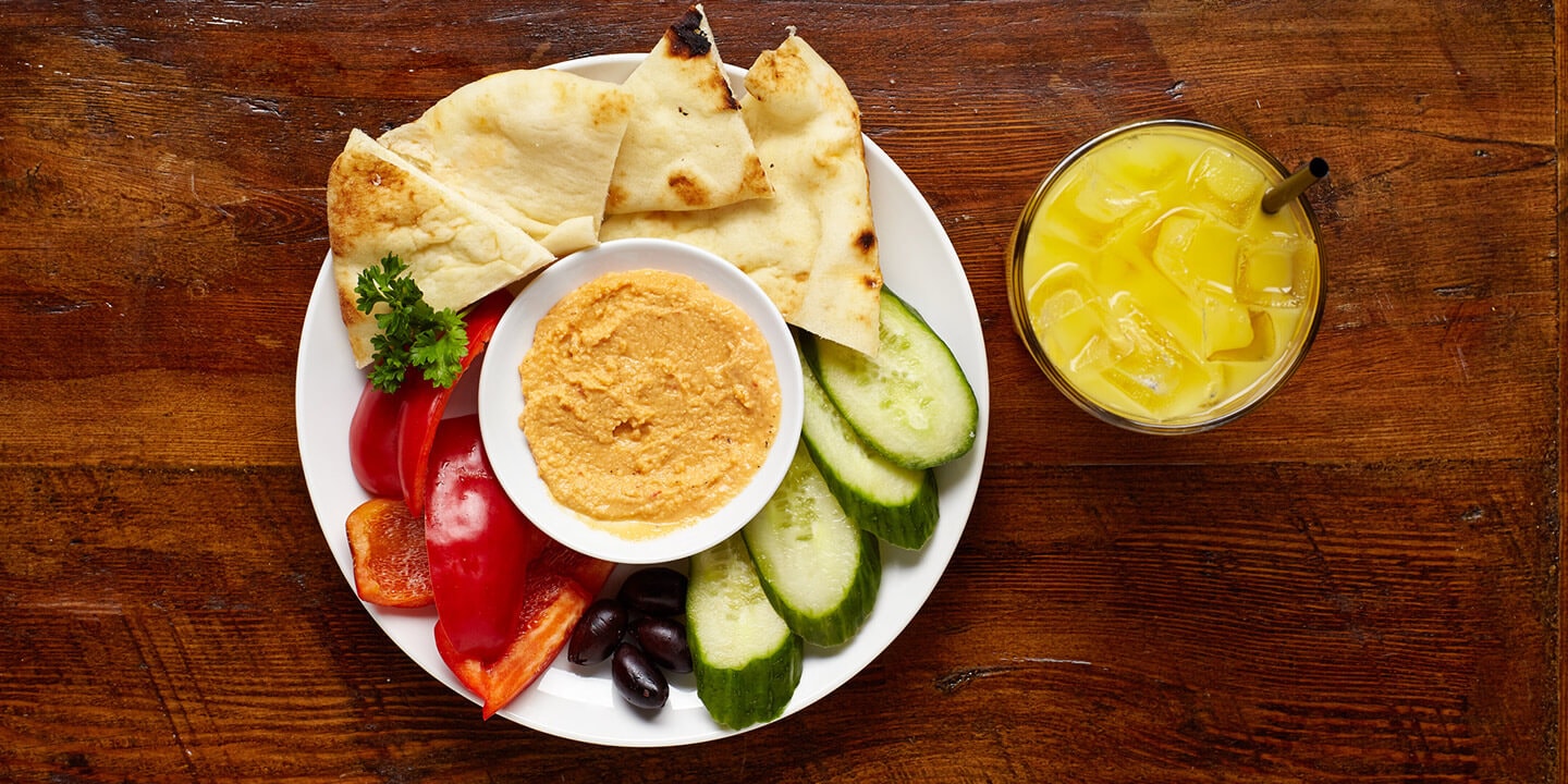 Bowl of hummus surrounded by olives, cucumbers, red peppers and pita bread, with a glass of orange juice